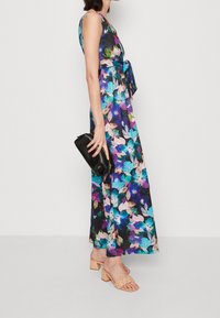 Robe maxi florale dans des couleurs sombres avec un bustier ajusté et une taille ceinturée. Une pochette noire et des sandales à talons carrés beige clair complètent la tenue.