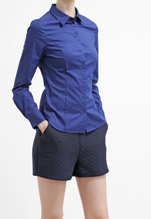 Frau trägt ein tailliertes blaues Hemd mit Knöpfen und gesteppten schwarzen Shorts, die Hände in den Taschen, und steht vor einem schlichten weißen Hintergrund.