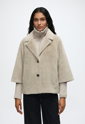 Vrouw met lang donker haar die een beige oversized pluizige jas draagt over een beige coltrui en zwarte broek, staand tegen een witte achtergrond.