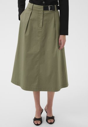 Kobieta w oliwkowozielonej spódnicy do kolan, czarnym pasku, czarnej bluzce z długim rękawem i czarnych butach na obcasie z odkrytymi palcami, stojąca na białej podłodze.