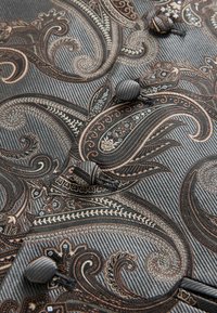 Teksturowana tkanina o szarym tle z misternymi wzorami paisley w brązowym i czarnym kolorze, podkreślona okrągłymi guzikami pokrytymi tkaniną.