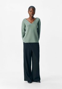 Vrouw die met handen achter de rug staat, gekleed in een saliegroene V-hals trui, wijde zwarte broek en zwarte schoenen op een witte achtergrond.
