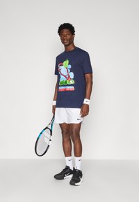 Granatowy t-shirt z grafiką, z motywem tenisowym, zestawiony z białymi szortami i czarnymi butami Nike. Trzyma rakietę tenisową z niebieskimi akcentami.