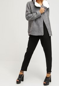 Grå jacka med dragkedja och vit pälskrage, tillsammans med svarta smala byxor och svarta platåklackar, som visar upp ett strukturerat tyg.