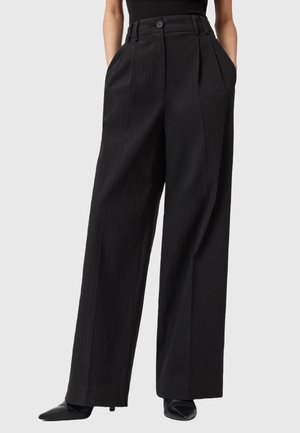 Persona che indossa pantaloni neri a gamba larga a vita alta con cintura con bottoni e mani nelle tasche, abbinati a scarpe nere con punta.