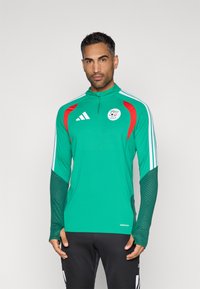 Zöld sportmez piros kiegészítőkkel, zippzáros gallérral és logóval. Texturált ujjakkal és fehér csíkokkal az oldalakon.