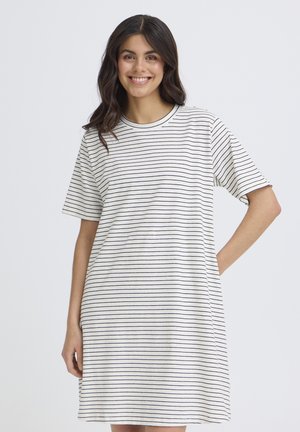 Noor naine naeratab, kandes valget lühikeste varrukatega kleiti, millel on õhukesed mustad horisontaalsed triibud, taust on ühevärviline.