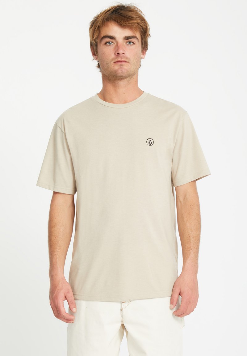 Beige katoenen T-shirt met korte mouwen en een ronde hals, met een klein zwart logo op de linkerborst, gladde textuur.