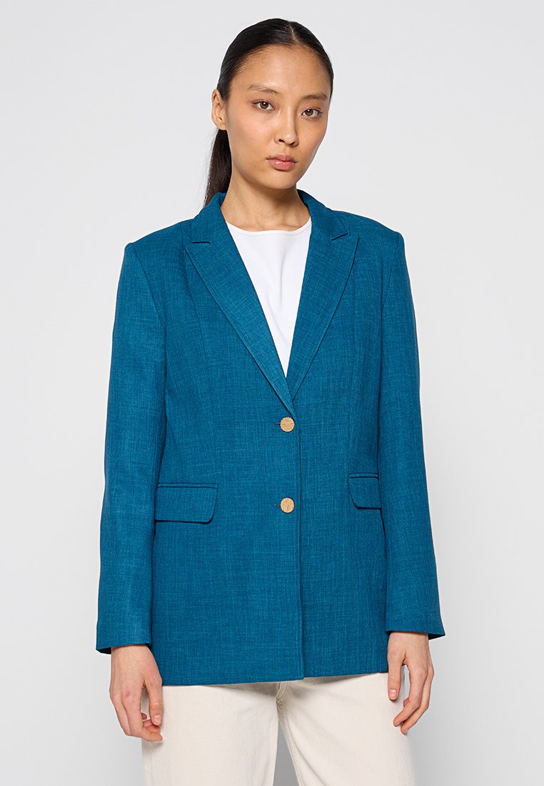 Blazer teal en tissu texturé, avec deux boutons dorés, des revers crantés et deux poches avant. Le modèle porte une chemise blanche en dessous.