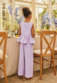 Orgonalila két részes öltözék ujjatlan, peplumos felsővel és bőszárú nadrággal, könnyű anyagból. Virágos asztal és fa székek mint háttér.