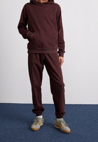 Bordová mikina s klokaní kapsou, doplněná o odpovídající bordové tepláky. Tkanina vypadá měkce. Oblečeno s šedými teniskami s vícebarevnými akcenty.