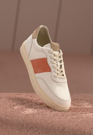 Baskets blanches et beige avec panneau latéral orange perforé, lacets beige et semelle beige sur une surface rose texturée.