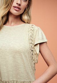 Jasnobeżowa bluzka z krótkimi rękawami i koronkowym detalem na ramieniu. Miękka struktura, luźny krój i falbaniasty dół.