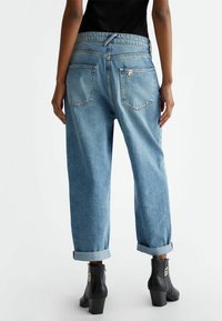 Henkilö, joka on pukeutunut löysästi istuviin, ylöspäin käärittyihin vaaleansinisiin farkkuihin ja mustiin korkokenkiin, seisoo tasaisella valkoisella taustalla.