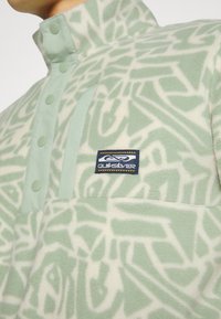 Pullover in felpa verde chiaro con motivi astratti bianchi, dotato di colletto alto, chiusura a bottone e patch con logo sul petto.