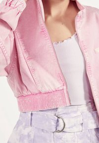 Kvinna som bär en ljusrosa kort jacka över en vit ribbad topp och högmidjade lavendelfärgade tie-dye byxor med en silverbältesspänne.