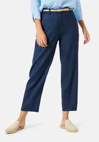 Námořnické modré lněné kalhoty s volným střihem, které mají světle béžový pásek, skládaný přední díl a boční kapsy; nošeny s béžovými botami na slip.