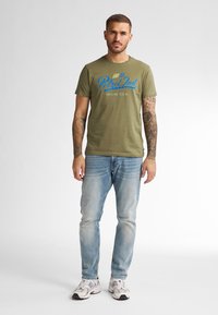 T-shirt en coton vert olive avec le texte graphique bleu "Petrol Ind" et "Miami U.S.A." ; associé à un jean bleu clair et des baskets blanches.
