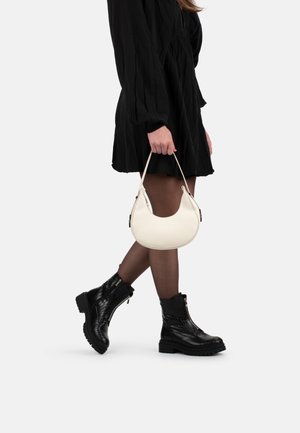 Personne portant une robe noire, des collants marron translucides et des bottines noires texturées, portant un petit sac à main blanc par sa poignée.
