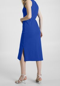 Mörkblå ärmlös klänning med figursydd bodice och slits bak, parat med vita remsandaler. Slät textur och knälång design.