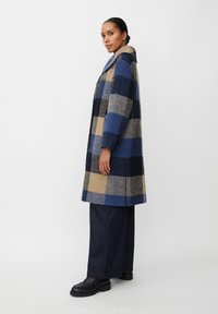 Rutet uldfrakke i nuancer af blå og beige, med krave og sideslids. Sæt sammen med vide denim-bukser og sorte klodset sko.