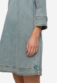 Světle modré džínové šaty s tříčtvrtečními rukávy, viditelným šitím a volným střihem. Mají rovný lem a mírně opotřebovaný vzhled.