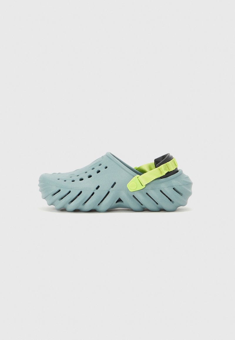 Light blue rubber clogs with ventilation holes, ridged sole design, adjustable green strap, and contrasting black toggle on the heel.