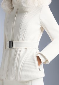 Jachetă albă matlasată cu glugă căptușită cu blană, curea în talie și buzunare cu fermoar. Prezintă nervuri texturate și material neted.