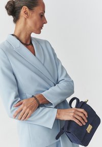 Lyseblå skræddersyet jakke med klapkrave, parret med en marineblå ruskinds håndtaske med en guldbuckle og korte håndtag, båret af en model.