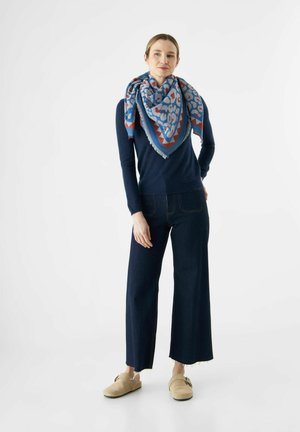 Une écharpe bleu à motifs drapée sur un pull navy, associée à un jean en denim foncé à jambe large et des chaussures beiges à enfiler avec un accent de boucle.