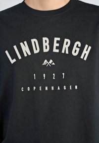 Camiseta de algodón negra con el texto en mayúsculas blancas "LINDBERGH 1927 COPENHAGUE" y un gráfico de montaña, cuello redondo y textura suave.