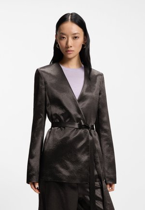 Vrouw met lang zwart haar draagt een glanzende zwarte blazer met ceintuur over een lichtpaarse top, staand tegen een effen witte achtergrond.