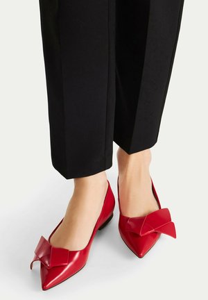 Stopy w czarnych spodniach i czerwonych balerinach z ostrym noskiem oraz dużymi ozdobnymi kokardami na górze, na jasnym tle.