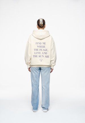 Personne portant un sweat à capuche crème avec le texte « Find me where the peace, love and the sun are » dans le dos, un jean bleu clair et des chaussures blanches, debout.