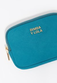 Bolso de tela en color verde azulado con el logo dorado "BIMBA Y LOLA", cierre con cremallera, forma rectangular, textura suave y detalle de costura en contraste.