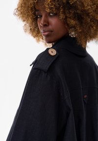 Czarna kurtka o strukturalnym designie, z wyraźnym kołnierzem, guzikiem na epauletach i teksturowanym wykończeniem. Zauważalny detal z guzikiem na ramieniu.