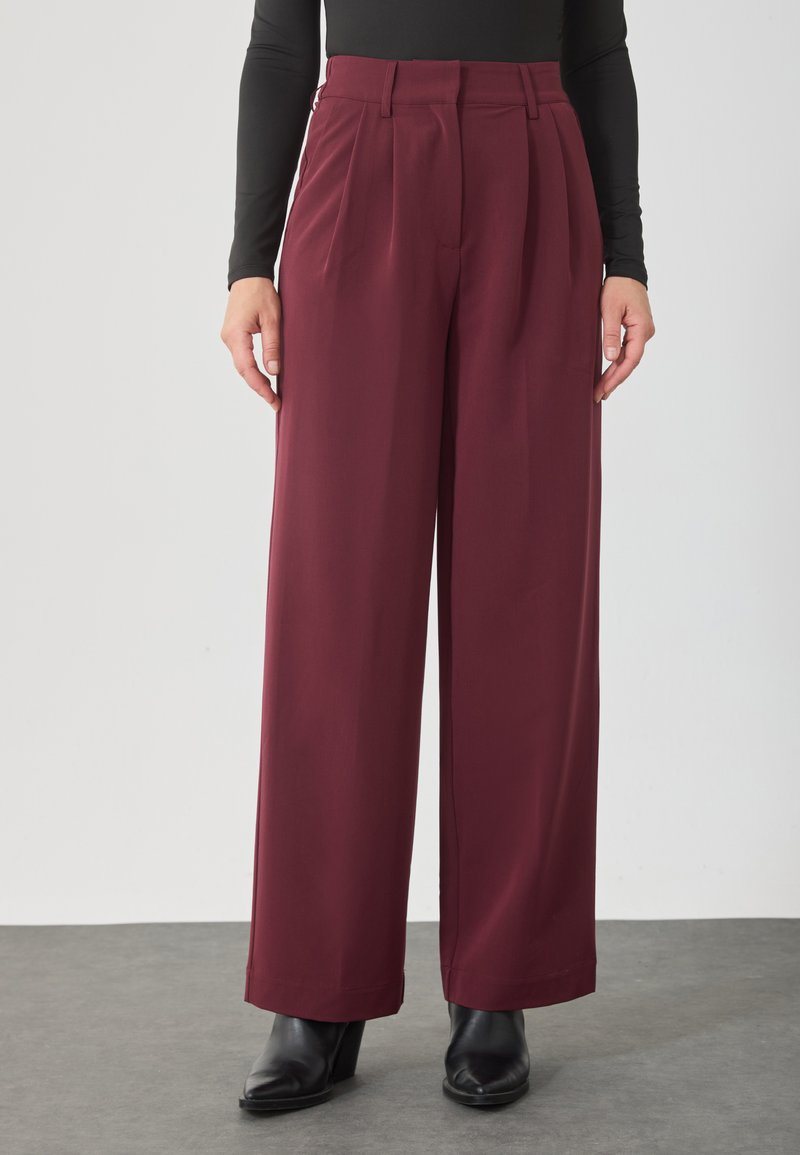 Osoba ubrana w bordowe, szerokie spodnie z wysokim stanem i plisami, w połączeniu z czarną bluzką oraz czarnymi botkami, stojąca na szarym podłożu.