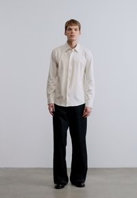 Απλό λευκό πουκάμισο με κουμπιά, συνδυασμένο με χαλαρά μαύρα παντελόνια. Λείο ύφασμα, απλός σχεδιασμός, χωρίς ορατά σχέδια ή διακοσμητικά στοιχεία.