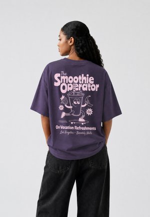 Kvinde med langt krøllet hår iført en lilla T-shirt viser rygprint af en smilende smoothie-kop på et skateboard med tekst om ferieopfriskninger.