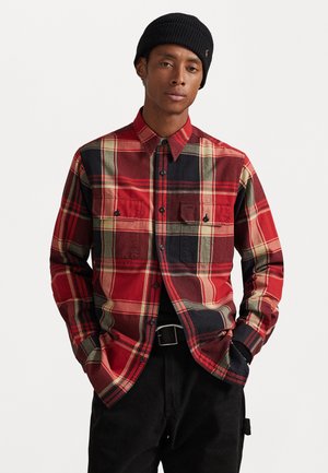 Red and black plaid flannel shirt with two chest pockets and black buttons. Long sleeves and a pointed collar, paired with black pants.