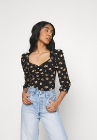 Sort blomstret bluse med lange ærmer, firkantet udskæring og sammensunket frontdetalje, parret med højtaljet lyseblå jeans.