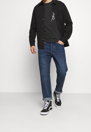 Mann trägt hochgekrempelte blaue Jeans, schwarze High-Top-Sneaker mit weißen Schnürsenkeln, schwarzes T-Shirt mit Grafik und unaufgeknöpftes schwarzes Hemd und läuft.