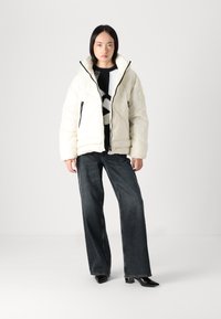 Jauna sieviete, ģērbusies baltā pild�ītā jakā, melnbaltā rakstainā džemperī, platās tumšās džinsās un melnās papēžu kurpēs, stāv balta fona priekšā.