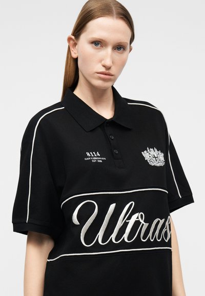 Νεαρή γυναίκα με ίσια, ανοιχτό καστανά μαλλιά φοράει μαύρο πόλο μπλουζάκι με λευκή ενίσχυση και κεντήματα "Ultras" και έμβλημα.