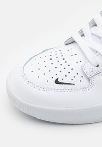 Sneaker bianca con design perforato e logo Nike nero. Presenta lacci piatti, suola in gomma e tomaia in sintetico liscio.