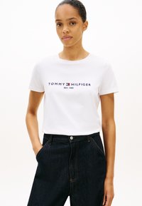 Biała bawełniana koszulka z krótkimi rękawami, z logo „TOMMY HILFIGER” w granatowym i czerwonym kolorze. Noszona z ciemnymi, wysokim stanem spodniami dżinsowymi.
