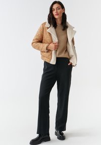 Beige gewatteerde jas met een witte fleece kraag, gedragen over een beige trui en zwarte broek; zwarte enkellaarsjes maken de outfit compleet.