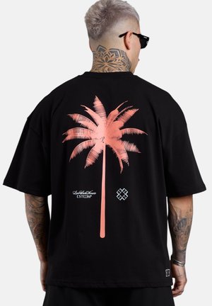 Person with tattooed neck and arms wears black oversized t-shirt with large pink palm tree graphic on the back.