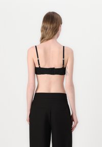 Čierny čipkový bralet s nastaviteľnými popruhmi, zapínaním na háčik a očko a hladkým zadným pásom. Modelka má oblečené čierne nohavice s vysokým pásom.