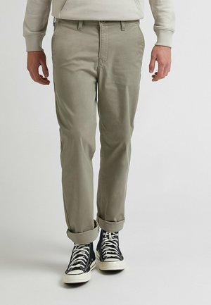 Man draagt beige broek met opgerolde pijpen, zwarte hoge sneakers met witte veters, en een lichtgekleurde trui.